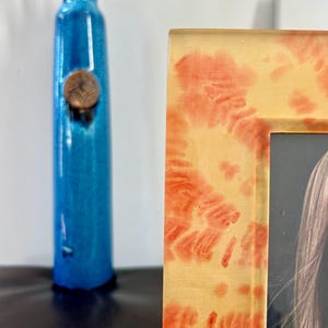 Op de afbeelding: Een hoge, turquoise blauwe vaas met een koperkleurig accent en een decoratieve fotolijst. De lijst heeft een gele achtergrond met rode en oranje patronen. De vaas bevat een paar bruine stelen.