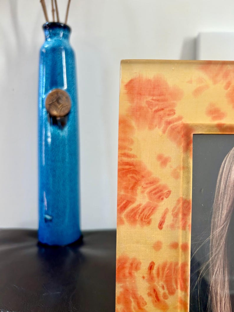 Op de afbeelding: Een hoge, turquoise blauwe vaas met een koperkleurig accent en een decoratieve fotolijst. De lijst heeft een gele achtergrond met rode en oranje patronen. De vaas bevat een paar bruine stelen.