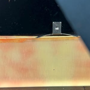Op de afbeelding: Close-up van een rechthoekig metalen stuk met een kleine schroef, bevestigd aan een goudkleurig oppervlak. Het oppervlak heeft een verloop van oranje en gele tinten. Een zwart oppervlak is zichtbaar op de achtergrond.