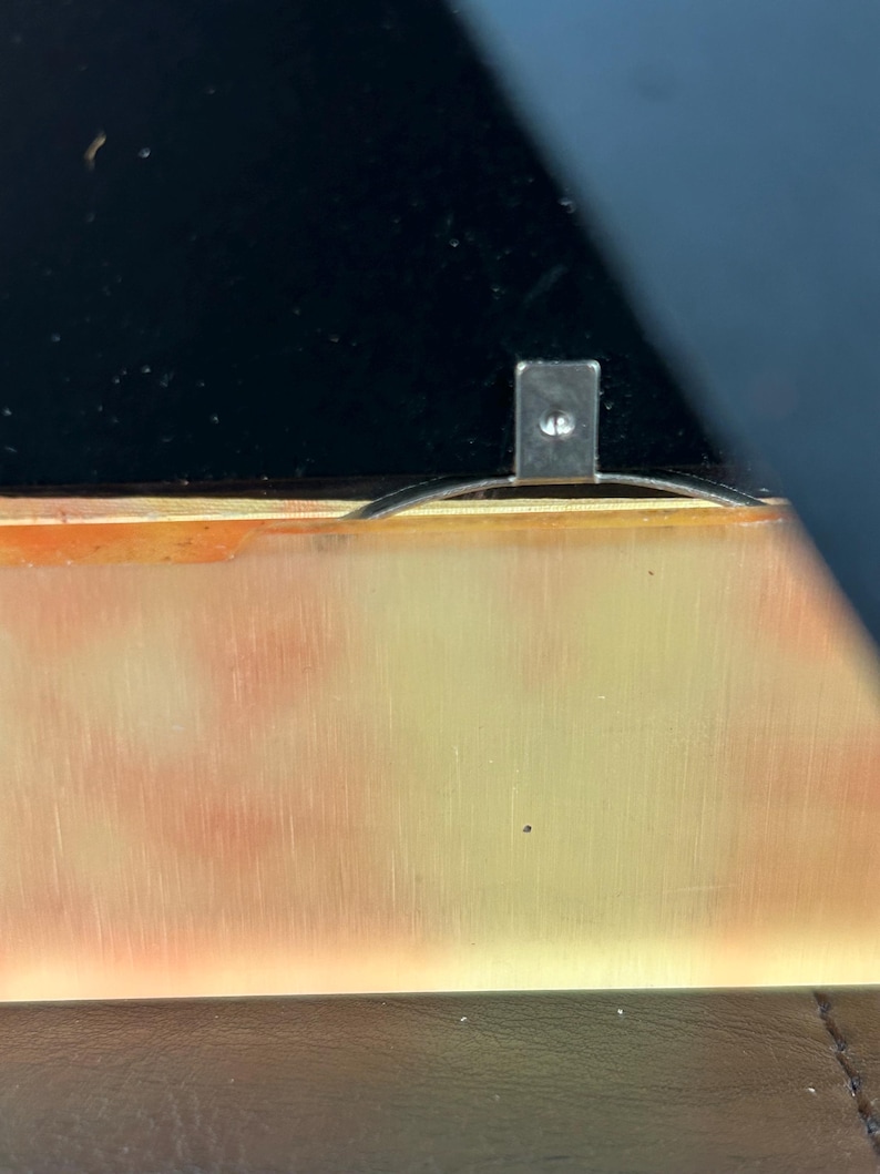Op de afbeelding: Close-up van een rechthoekig metalen stuk met een kleine schroef, bevestigd aan een goudkleurig oppervlak. Het oppervlak heeft een verloop van oranje en gele tinten. Een zwart oppervlak is zichtbaar op de achtergrond.
