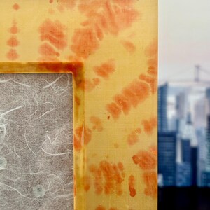 Op de afbeelding: Close-up van een rechthoekige fotolijst met een geel en oranje tie-dye patroon. De lijst heeft een textuur en een neutrale achtergrond. De achtergrond toont een vage stadsgezicht.