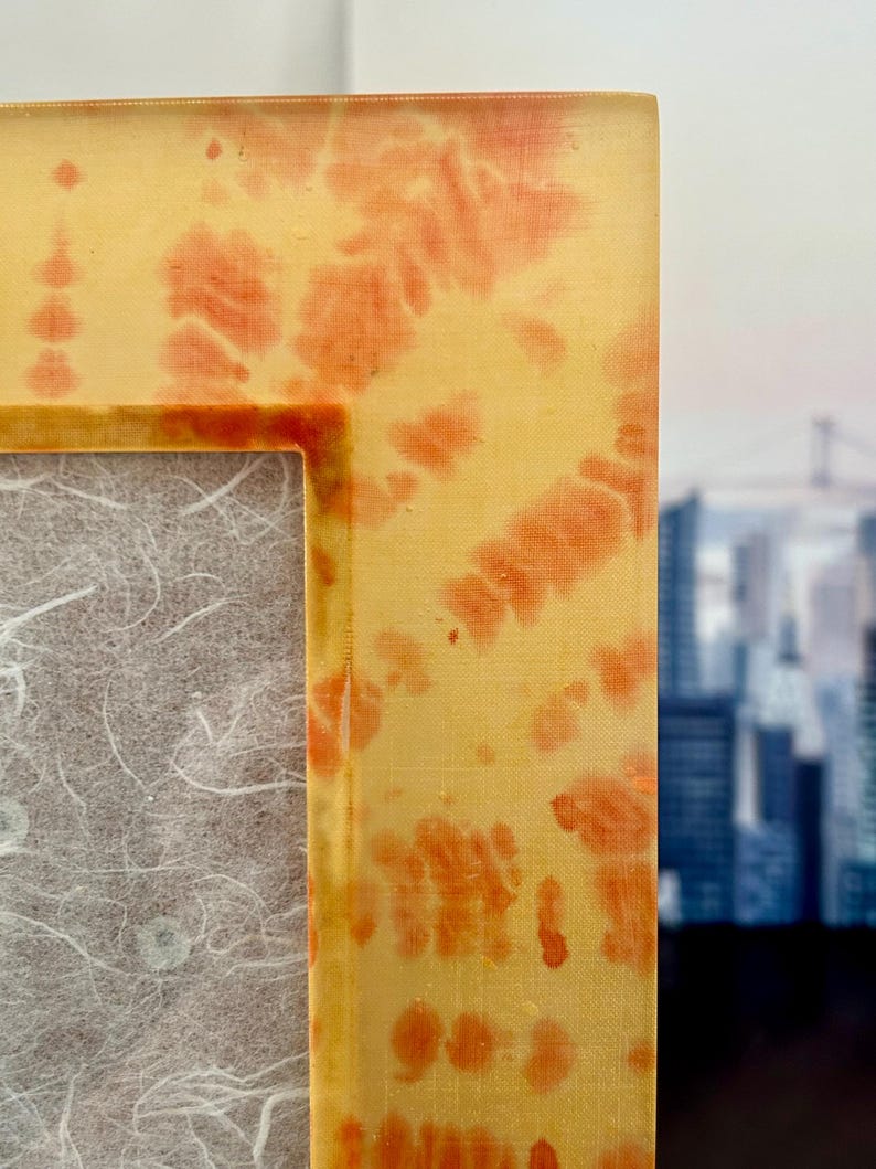 Op de afbeelding: Close-up van een rechthoekige fotolijst met een geel en oranje tie-dye patroon. De lijst heeft een textuur en een neutrale achtergrond. De achtergrond toont een vage stadsgezicht.