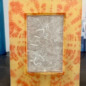Op de afbeelding: Een rechthoekige fotolijst met een geel en oranje tie-dye patroon. De lijst heeft een heldere, glanzende afwerking en een rechthoekige opening voor een foto. De achtergrond is een getextureerd, gebroken wit papier.