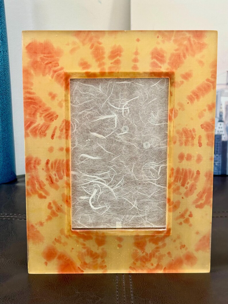 Op de afbeelding: Een rechthoekige fotolijst met een geel en oranje tie-dye patroon. De lijst heeft een heldere, glanzende afwerking en een rechthoekige opening voor een foto. De achtergrond is een getextureerd, gebroken wit papier.