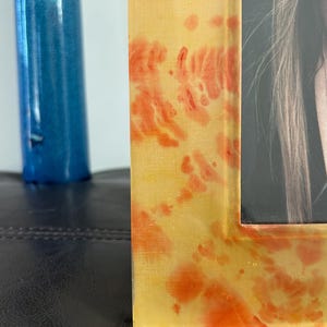 Op de afbeelding: Close-up van een rechthoekige fotolijst met een geel en oranje gevlekt ontwerp. De lijst bevat een zwart-witfoto. Een blauw decoratief object is gedeeltelijk zichtbaar op de achtergrond.