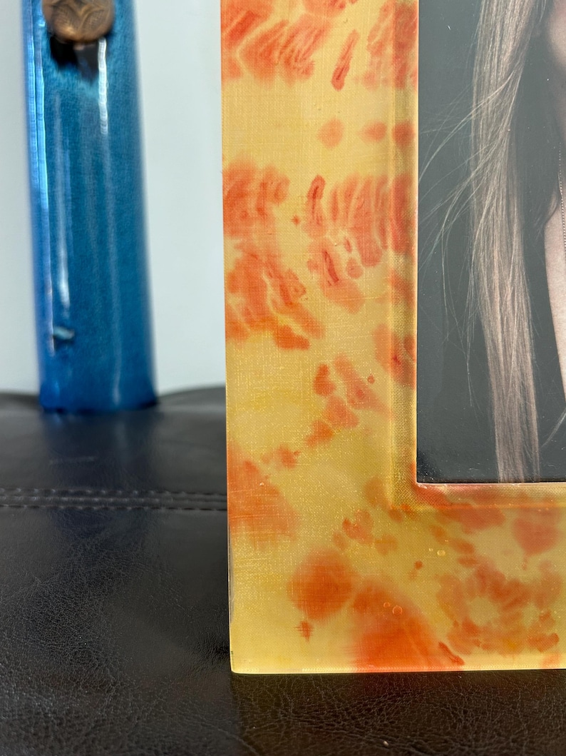 Op de afbeelding: Close-up van een rechthoekige fotolijst met een geel en oranje gevlekt ontwerp. De lijst bevat een zwart-witfoto. Een blauw decoratief object is gedeeltelijk zichtbaar op de achtergrond.