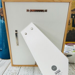 May include: Back of a wooden picture frame with a white backing and a light brown wooden frame. The frame has a metal sawtooth hanger and a metal bracket for hanging. A white easel back is attached to the frame.