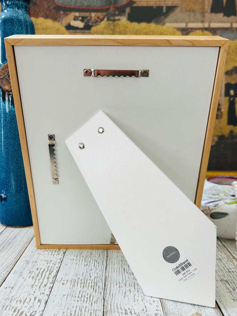 May include: Back of a wooden picture frame with a white backing and a light brown wooden frame. The frame has a metal sawtooth hanger and a metal bracket for hanging. A white easel back is attached to the frame.