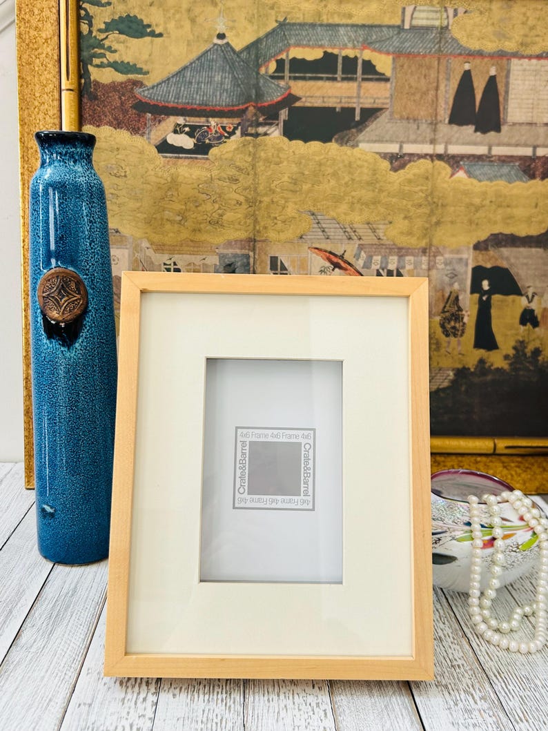 May include: A rectangular wooden picture frame with a cream-colored mat and a clear glass cover. The frame is labeled "Crate & Barrel" and is designed for a 4x6 inch photo. A blue vase and a decorative painting are in the background.