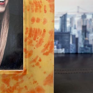Op de afbeelding: Een close-up van een fotolijst met een geel en oranje tie-dye patroon. De lijst is gemaakt van een doorschijnend materiaal en heeft een glanzende afwerking. De foto in de lijst is gedeeltelijk zichtbaar.