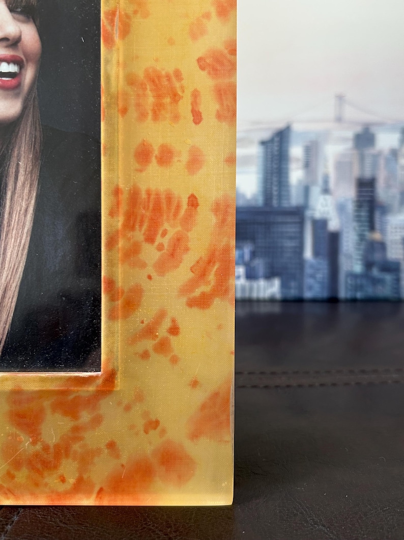 Op de afbeelding: Een close-up van een fotolijst met een geel en oranje tie-dye patroon. De lijst is gemaakt van een doorschijnend materiaal en heeft een glanzende afwerking. De foto in de lijst is gedeeltelijk zichtbaar.