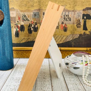 May include: A light-colored wooden picture frame with a white backing, standing on a white-washed wooden surface. The frame is angled, showcasing its construction. A blue vase and decorative items are in the background.