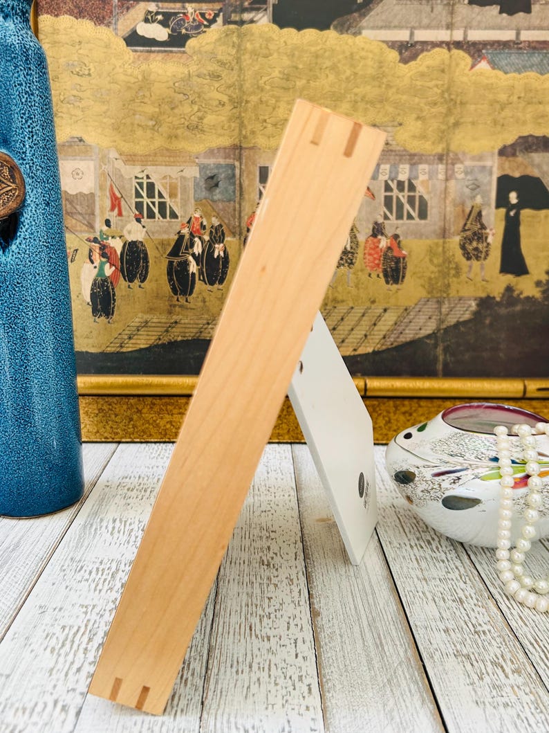 May include: A light-colored wooden picture frame with a white backing, standing on a white-washed wooden surface. The frame is angled, showcasing its construction. A blue vase and decorative items are in the background.