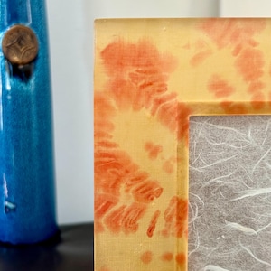 Op de afbeelding: Close-up van een fotolijst met een geel en oranje tie-dye patroon. De lijst heeft een rechthoekige opening en een getextureerde, gebroken witte achterkant. Een blauwe vaas met een bronzen stop is op de achtergrond.