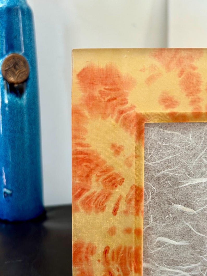 Op de afbeelding: Close-up van een fotolijst met een geel en oranje tie-dye patroon. De lijst heeft een rechthoekige opening en een getextureerde, gebroken witte achterkant. Een blauwe vaas met een bronzen stop is op de achtergrond.