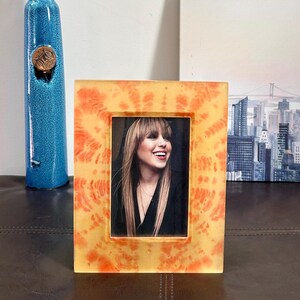 Op de afbeelding: Een rechthoekige fotolijst met een oranje en geel tie-dye patroon. De lijst bevat een zwart-wit foto van een lachende persoon. Links staat een hoge blauwe vaas met een decoratieve bovenkant.