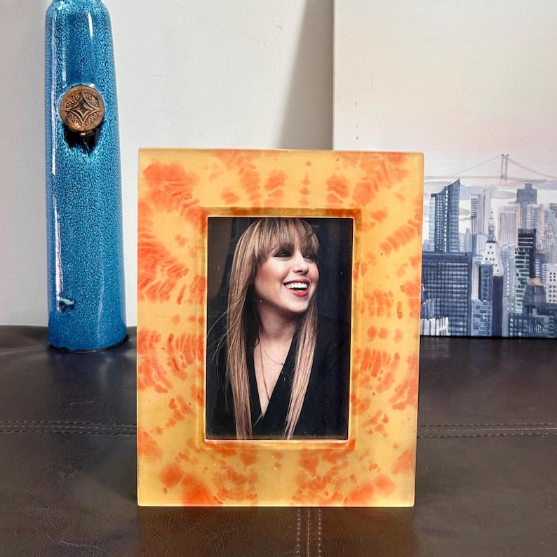 Op de afbeelding: Een rechthoekige fotolijst met een oranje en geel tie-dye patroon. De lijst bevat een zwart-wit foto van een lachende persoon. Links staat een hoge blauwe vaas met een decoratieve bovenkant.