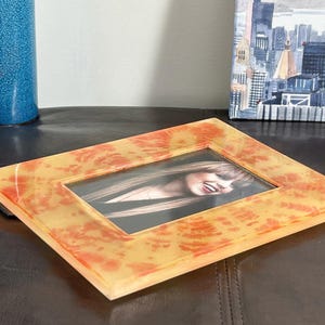Op de afbeelding: Een rechthoekige fotolijst met een geel en oranje gemarmerd ontwerp. De lijst bevat een foto van een lachende persoon. De lijst is gemaakt van een doorschijnend materiaal en ligt op een donkerbruin oppervlak.