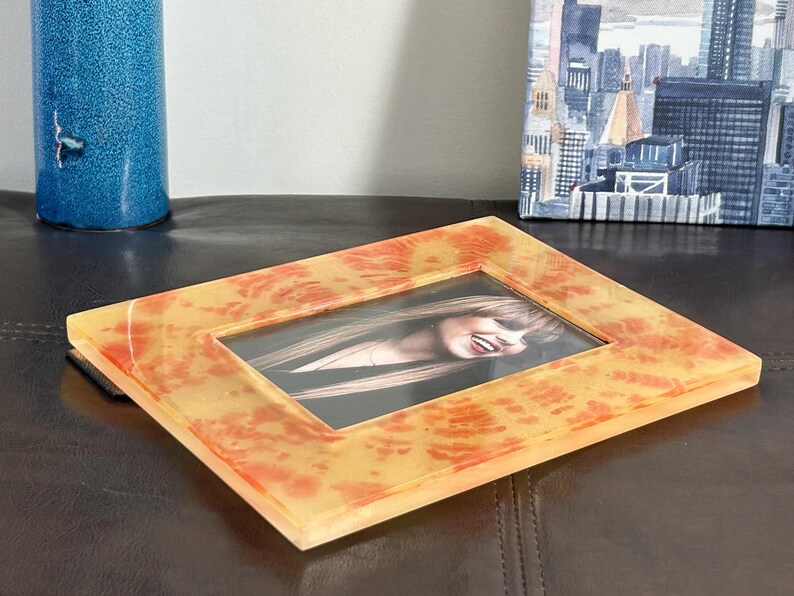 Op de afbeelding: Een rechthoekige fotolijst met een geel en oranje gemarmerd ontwerp. De lijst bevat een foto van een lachende persoon. De lijst is gemaakt van een doorschijnend materiaal en ligt op een donkerbruin oppervlak.