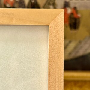 May include: Close-up of a light-colored wooden picture frame with a light cream-colored matte. The frame has a simple, clean design with a natural wood grain. The background shows a glimpse of a painting.