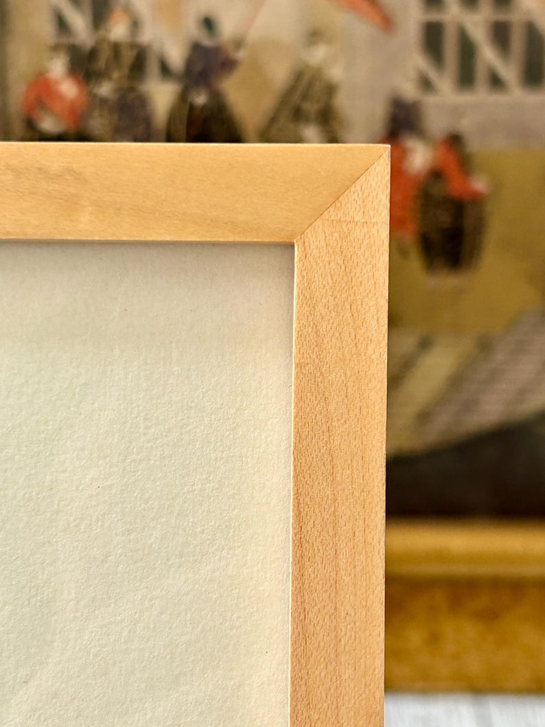 May include: Close-up of a light-colored wooden picture frame with a light cream-colored matte. The frame has a simple, clean design with a natural wood grain. The background shows a glimpse of a painting.