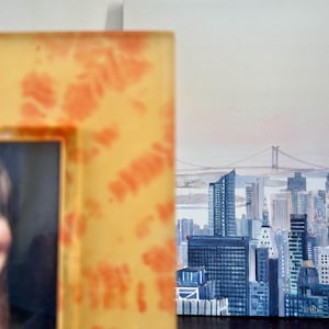 Op de afbeelding: Een ingelijst schilderij van een stadsgezicht, met de skyline van New York City met wolkenkrabbers en een brug. De lijst is geel met oranje tie-dye patronen. Het schilderij staat op een wit canvas.