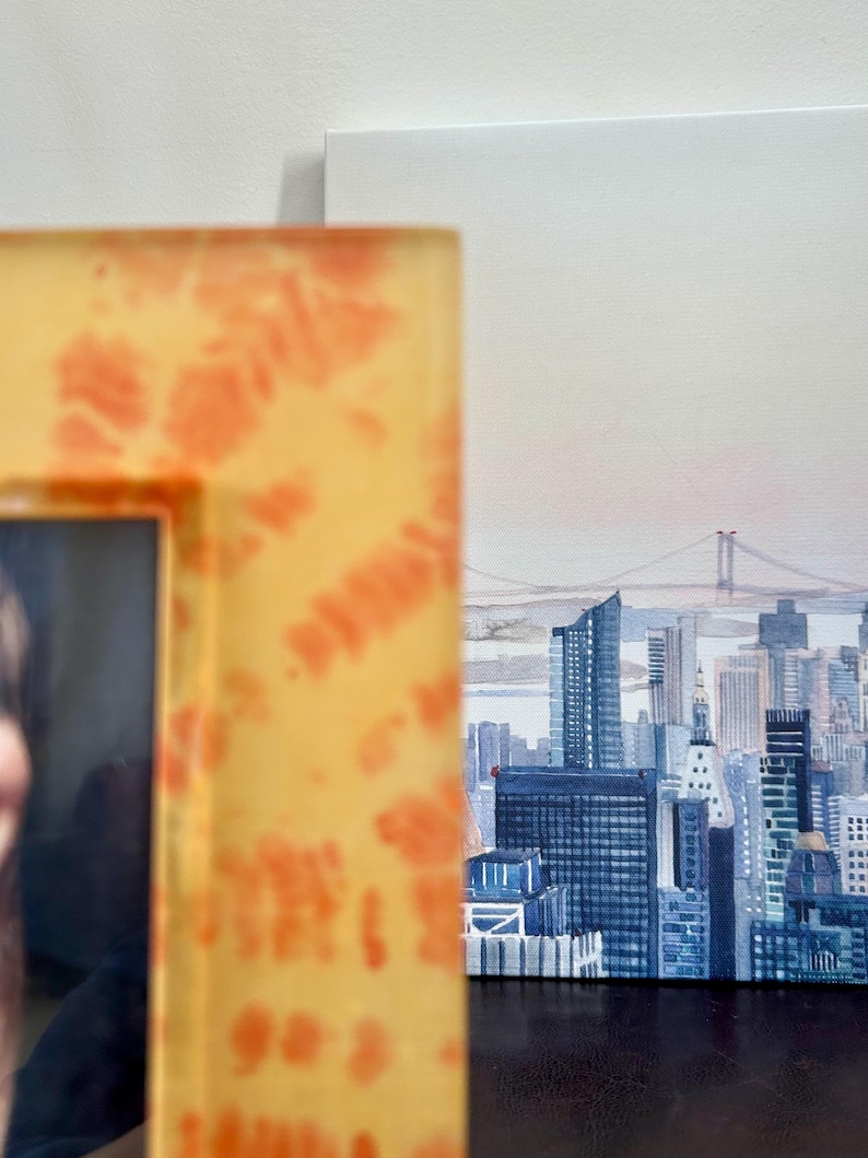 Op de afbeelding: Een ingelijst schilderij van een stadsgezicht, met de skyline van New York City met wolkenkrabbers en een brug. De lijst is geel met oranje tie-dye patronen. Het schilderij staat op een wit canvas.