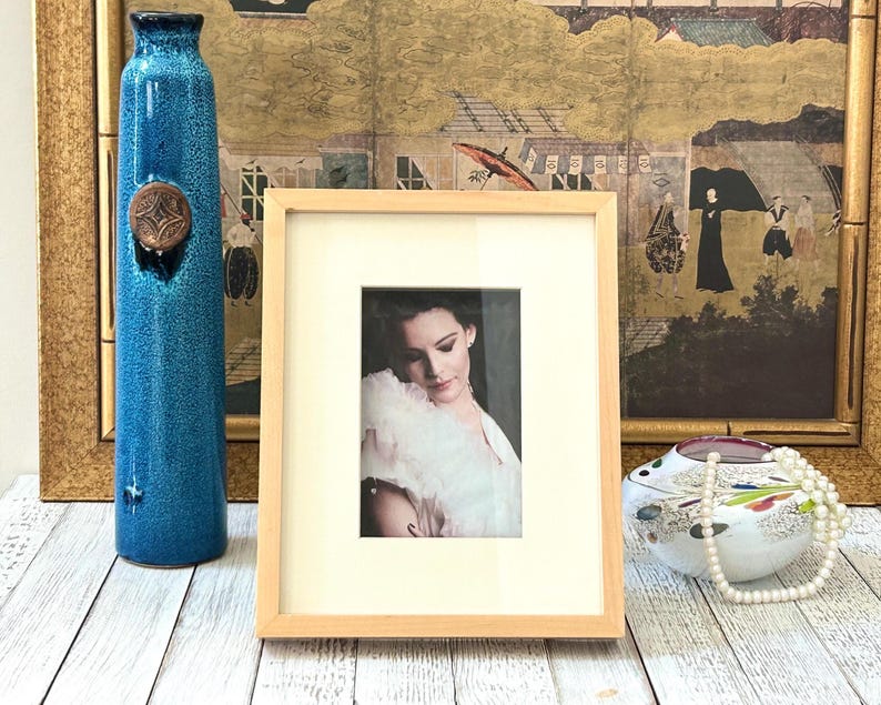 May include: A tall, turquoise ceramic vase with a copper-colored accent sits next to a framed portrait. A decorative, light-colored wooden frame surrounds the portrait of a woman. A decorative, white and multi-colored ceramic bowl with a pearl necklace is also present.