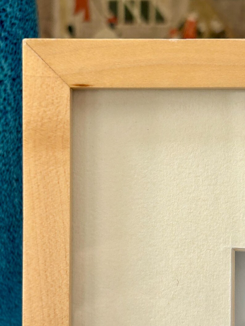 May include: A close-up of a light-colored wooden picture frame with a white mat. The frame has a simple, clean design with a natural wood grain finish. The corner of the frame is visible, showing the precise joinery. The background is a textured, off-white surface.