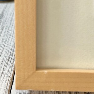 May include: A close-up of a light-colored wooden picture frame. The frame has a simple, rectangular design with a natural wood finish. The frame surrounds a white or off-white surface, possibly a print or artwork.