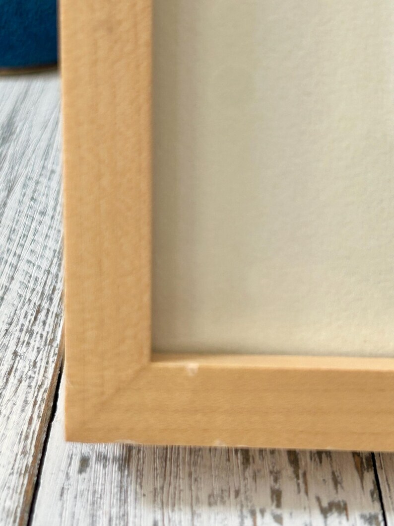 May include: A close-up of a light-colored wooden picture frame. The frame has a simple, rectangular design with a natural wood finish. The frame surrounds a white or off-white surface, possibly a print or artwork.