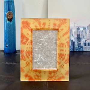 Op de afbeelding: Een rechthoekige fotolijst met een tie-dye patroon in geel- en oranjetinten. Het frame bevat een getextureerd, gebroken wit papier. Links is een blauwe vaas met een bronzen embleem te zien.