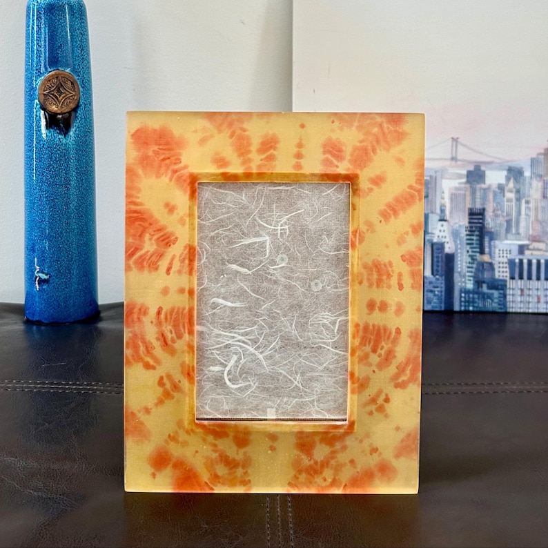Op de afbeelding: Een rechthoekige fotolijst met een tie-dye patroon in geel- en oranjetinten. Het frame bevat een getextureerd, gebroken wit papier. Links is een blauwe vaas met een bronzen embleem te zien.