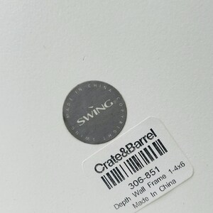 May include: A close-up of a white surface with two labels. One label is a gray circle with the text "MADE IN CHINA SWING COPYRIGHT". The other label is a white rectangle with the text "Crate & Barrel" and "Depth Wall Frame 1-4x6 Made In China".