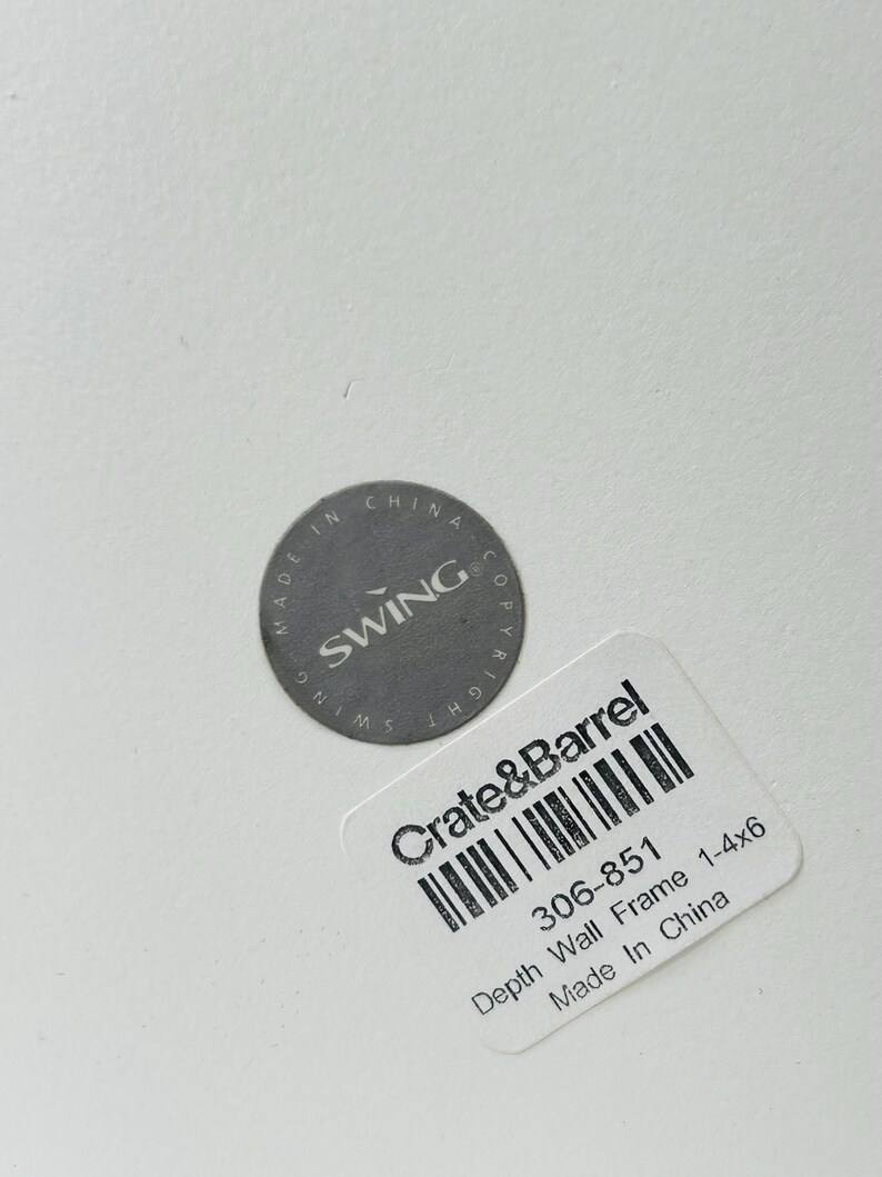 May include: A close-up of a white surface with two labels. One label is a gray circle with the text "MADE IN CHINA SWING COPYRIGHT". The other label is a white rectangle with the text "Crate & Barrel" and "Depth Wall Frame 1-4x6 Made In China".