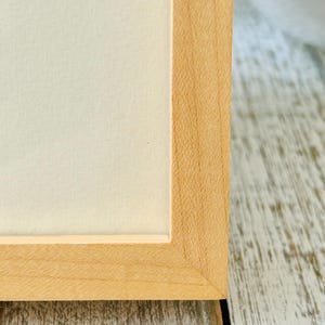 May include: Close-up of a light-colored wooden picture frame with a white interior. The frame has a natural wood grain and a simple, clean design. The frame is sitting on a distressed, light-colored wooden surface.