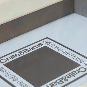 May include: A close-up of a gray wooden picture frame with a white interior. The frame has a dark gray border and the text "Crate & Barrel" and "4x6 Frame" are printed on the white interior.