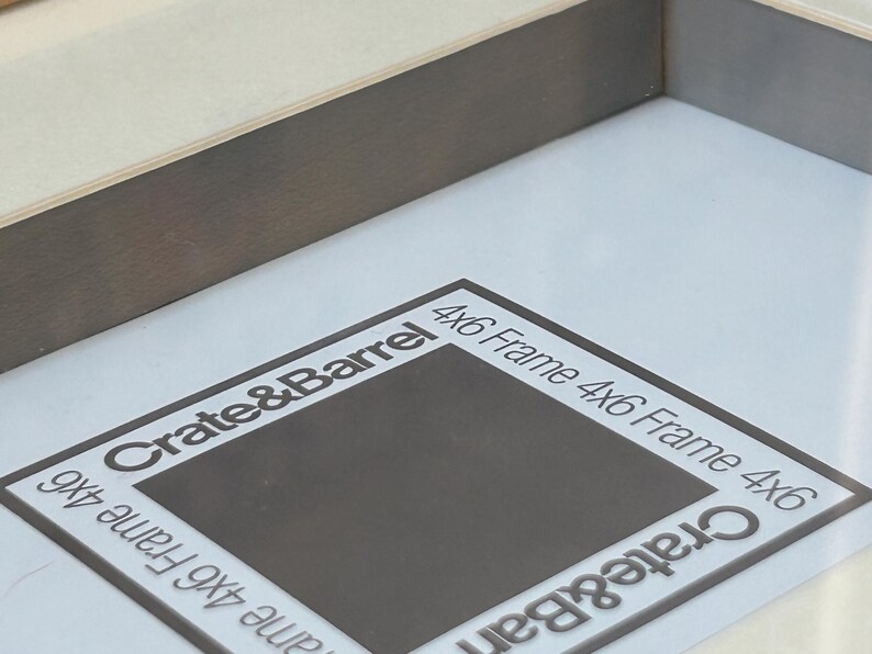 May include: A close-up of a gray wooden picture frame with a white interior. The frame has a dark gray border and the text "Crate & Barrel" and "4x6 Frame" are printed on the white interior.