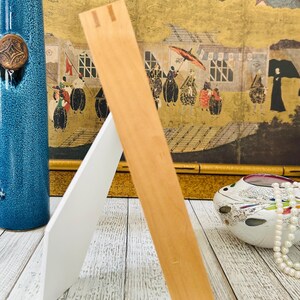 May include: A wooden picture frame with a natural finish, propped up on a white backing. The frame is angled, showcasing its construction. A blue vase and decorative items are in the background.