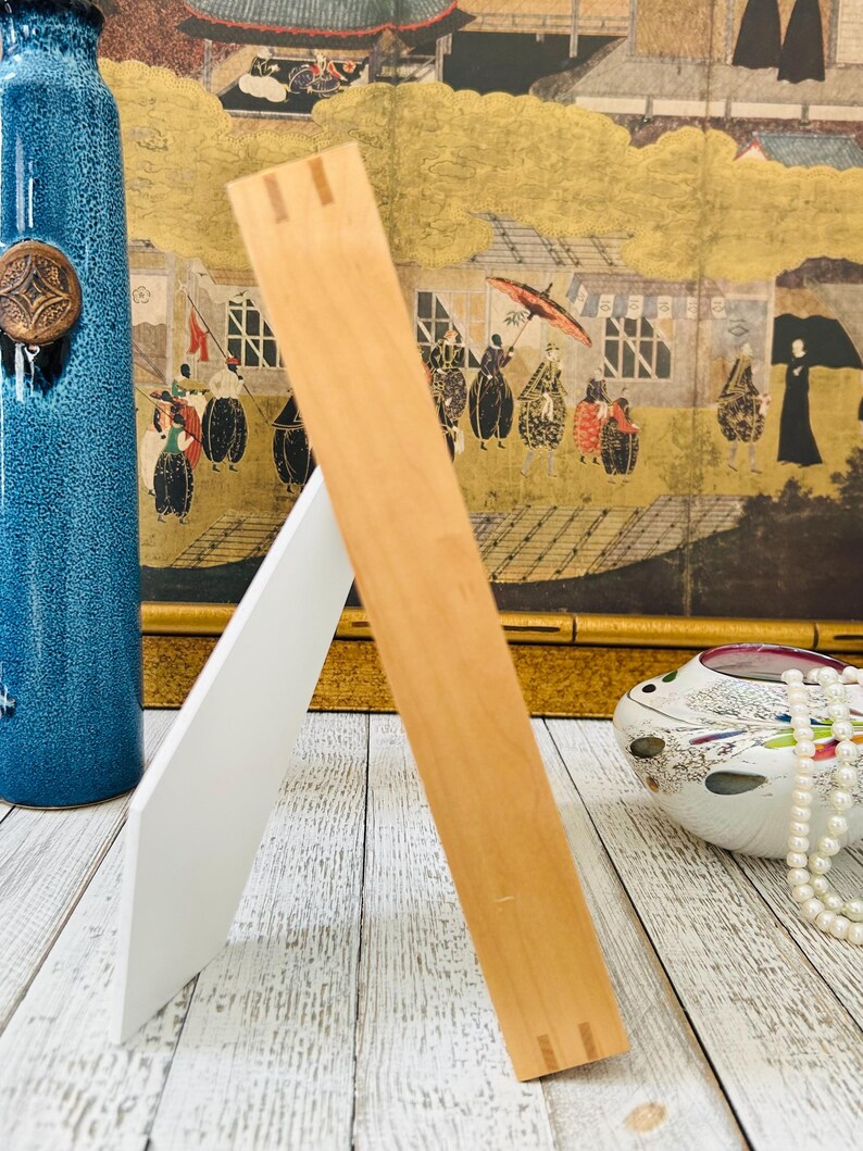 May include: A wooden picture frame with a natural finish, propped up on a white backing. The frame is angled, showcasing its construction. A blue vase and decorative items are in the background.