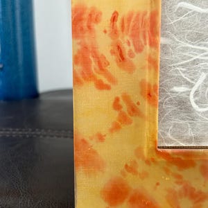 Op de afbeelding: Een close-up van een decoratieve fotolijst met een geel en oranje gemarmerd ontwerp. De lijst heeft een rechthoekige vorm en een gestructureerd oppervlak. De lijst staat naast een blauw cilindrisch object.