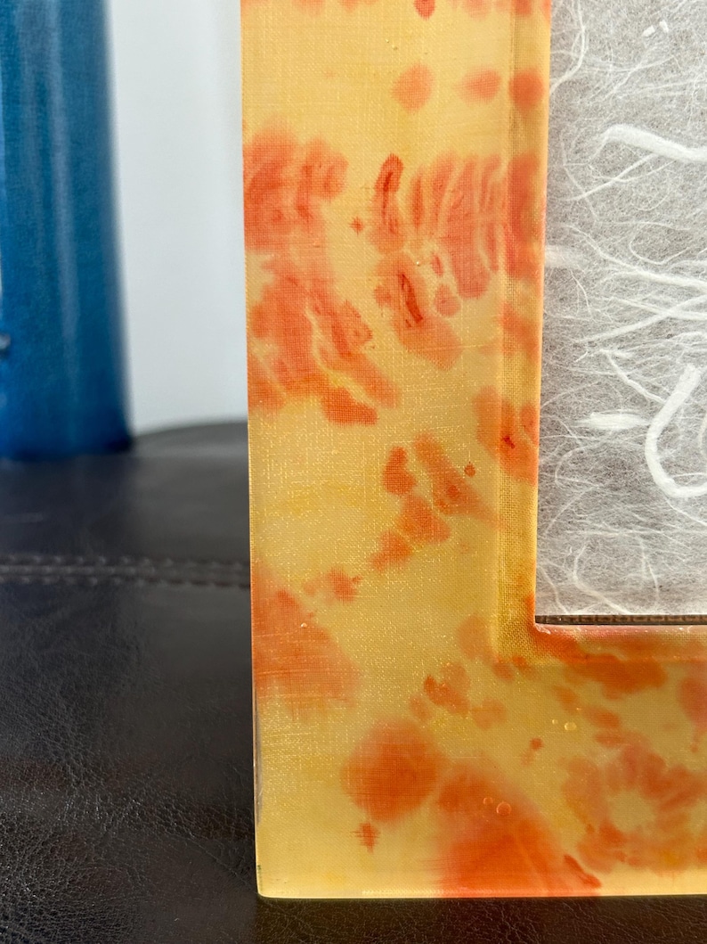 Op de afbeelding: Een close-up van een decoratieve fotolijst met een geel en oranje gemarmerd ontwerp. De lijst heeft een rechthoekige vorm en een gestructureerd oppervlak. De lijst staat naast een blauw cilindrisch object.