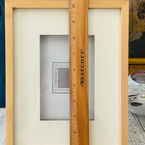 May include: A light-colored wooden picture frame with a white mat, designed for a 4x6 inch photo. A wooden ruler, marked with inches, is placed vertically in front of the frame, showing measurements.
