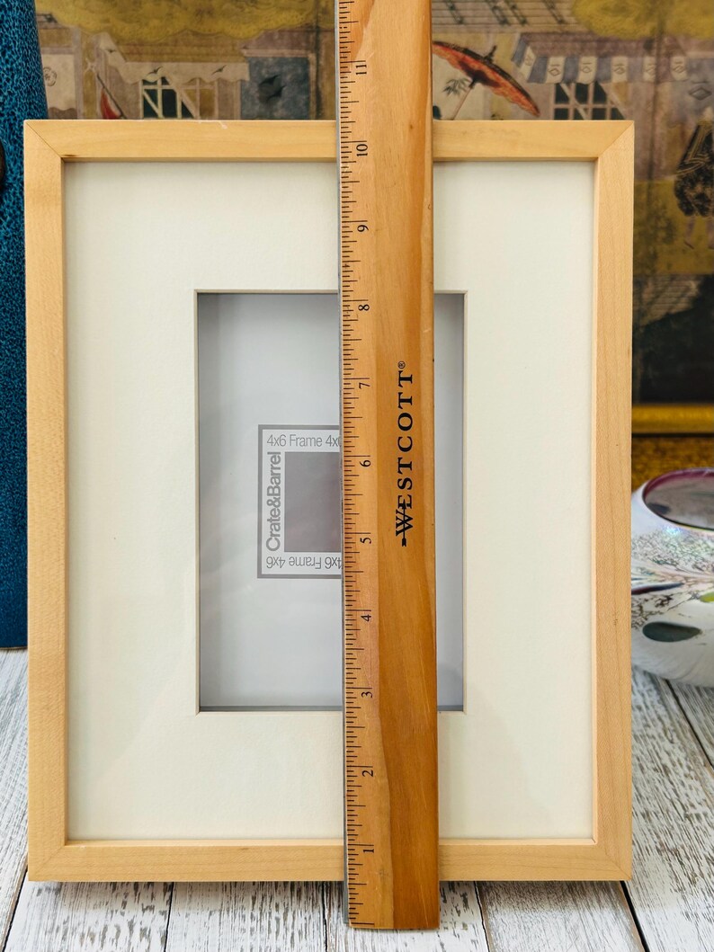 May include: A light-colored wooden picture frame with a white mat, designed for a 4x6 inch photo. A wooden ruler, marked with inches, is placed vertically in front of the frame, showing measurements.