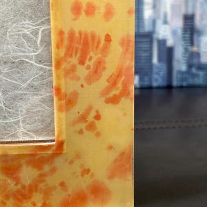 Op de afbeelding: Close-up van een rechthoekige fotolijst met een geel en oranje tie-dye patroon. De lijst heeft een heldere, glanzende afwerking en een witte, getextureerde achterkant. De achtergrond is wazig en toont een stadsgezicht.