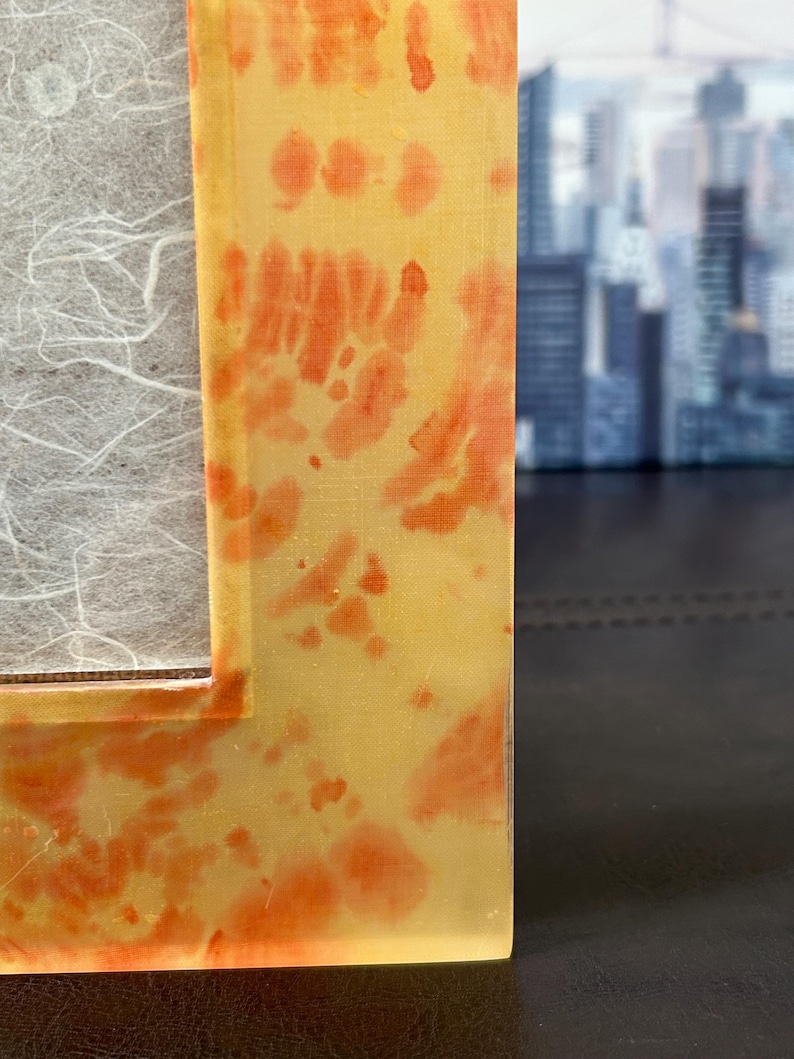 Op de afbeelding: Close-up van een rechthoekige fotolijst met een geel en oranje tie-dye patroon. De lijst heeft een heldere, glanzende afwerking en een witte, getextureerde achterkant. De achtergrond is wazig en toont een stadsgezicht.