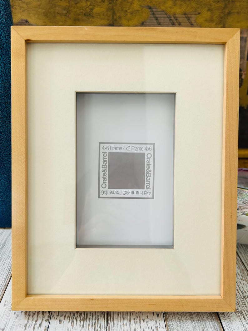 May include: A rectangular wooden picture frame with a cream-colored mat. The frame is light brown and has the text "4x6 Frame 4x6 Frame 4x6" and "Crate&Barrel" printed on the mat.