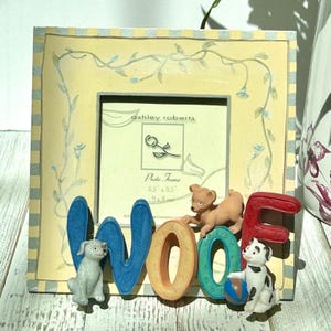 May include: A decorative photo frame with the word "WOOF" spelled out in colorful letters and dog figurines. The frame is yellow with a floral design and the text "ashley roberts" and "Photo Frame" are visible.
