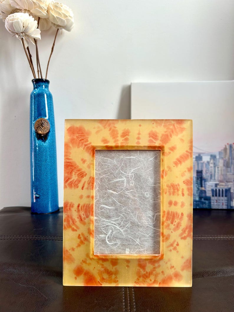 Op de afbeelding: Een rechthoekige fotolijst met een tie-dye ontwerp in oranje en geel. De lijst heeft een heldere, glanzende afwerking en een witte papieren achterkant. Een blauwe vaas met witte bloemen staat op de achtergrond.