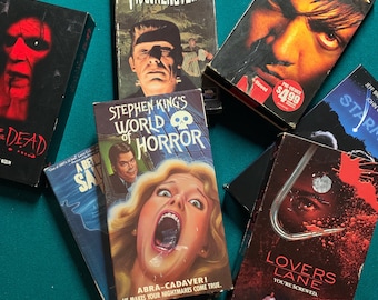 May include: A collection of VHS tapes featuring horror movies. The tapes are stacked on a green felt surface. The titles include "House of Frankenstein", "Stephen King's World of Horror", "Dahmer", "Lovers Lane", and "The Dead".
